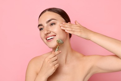 Beautiful young woman doing facial massage with roller on pink background