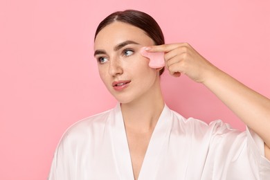 Photo of Beautiful young woman doing facial massage with gua sha tool on pink background