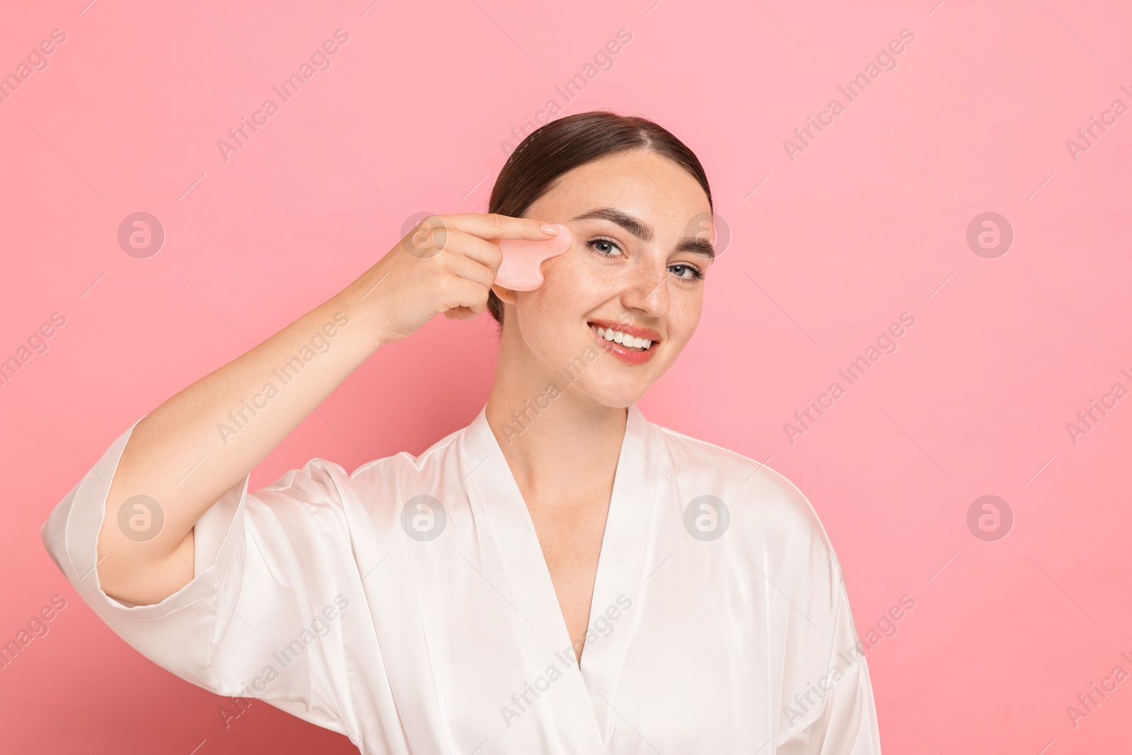 Photo of Beautiful young woman doing facial massage with gua sha tool on pink background