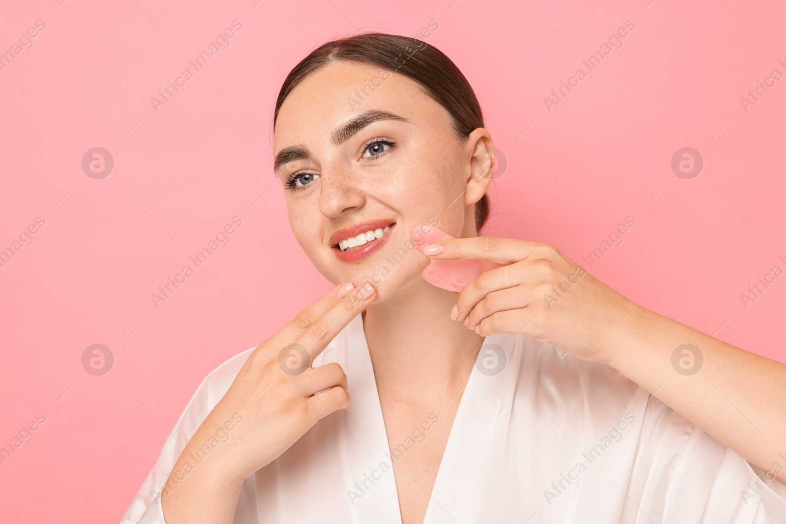 Photo of Beautiful young woman doing facial massage with gua sha tool on pink background