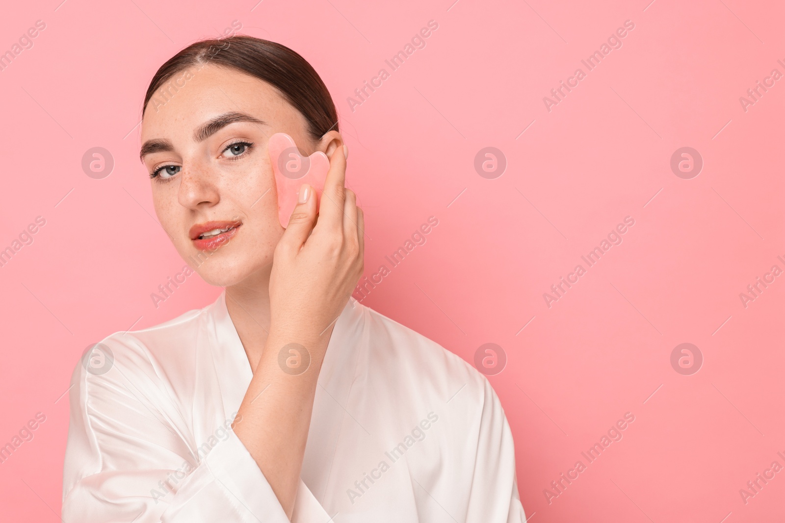 Photo of Beautiful young woman doing facial massage with gua sha tool on pink background, space for text