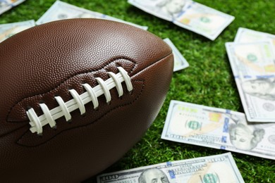 Photo of American football ball and dollar banknotes on green grass outdoors, closeup