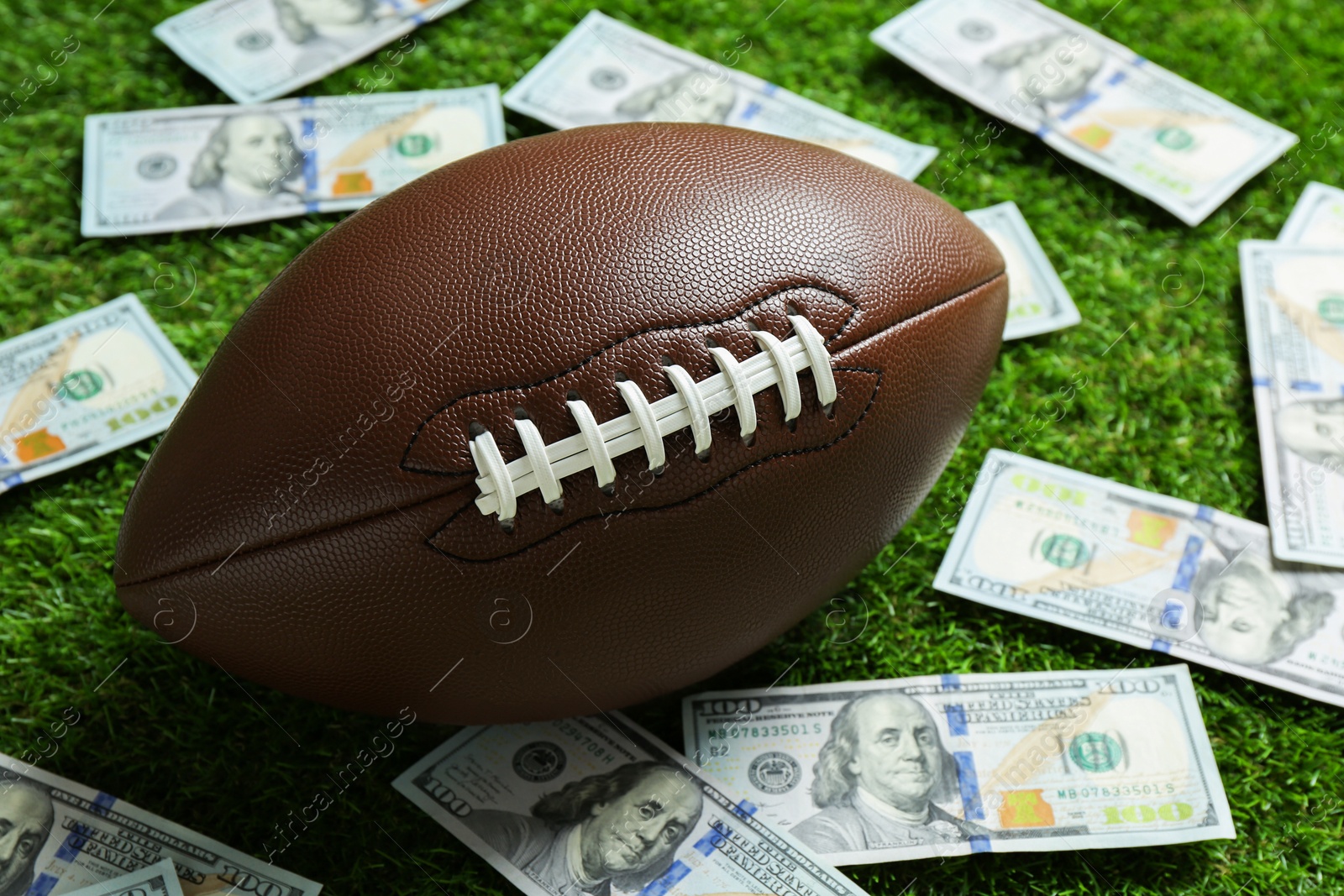 Photo of American football ball and dollar banknotes on green grass outdoors, closeup