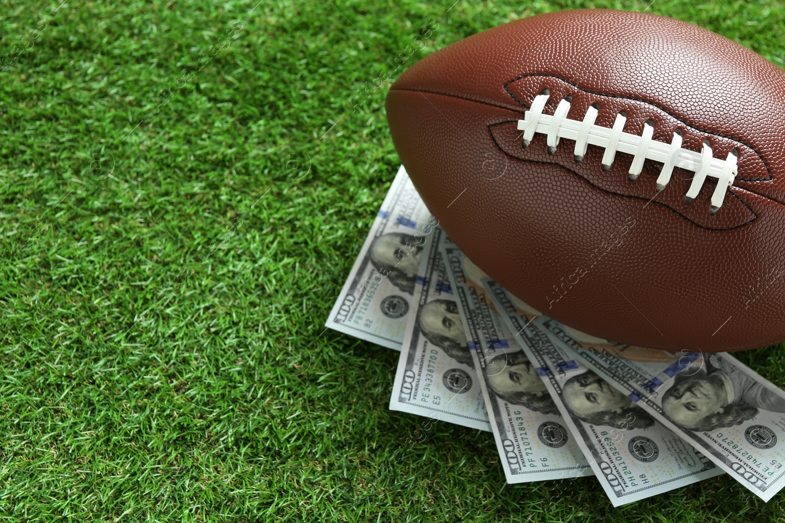 Photo of American football ball and dollar banknotes on green grass outdoors, space for text