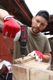 Photo of Man twisting screw into wooden plank with electric screwdriver outdoors, selective focus