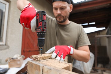 Photo of Man twisting screw into wooden plank with electric screwdriver outdoors, selective focus