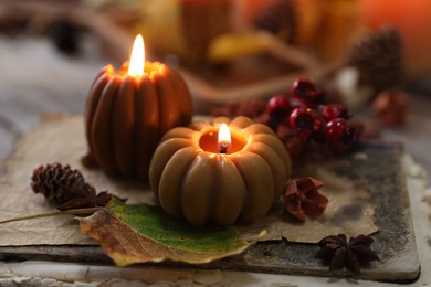 Photo of Burning candles in shape of pumpkins, old book and autumn decor on table, closeup