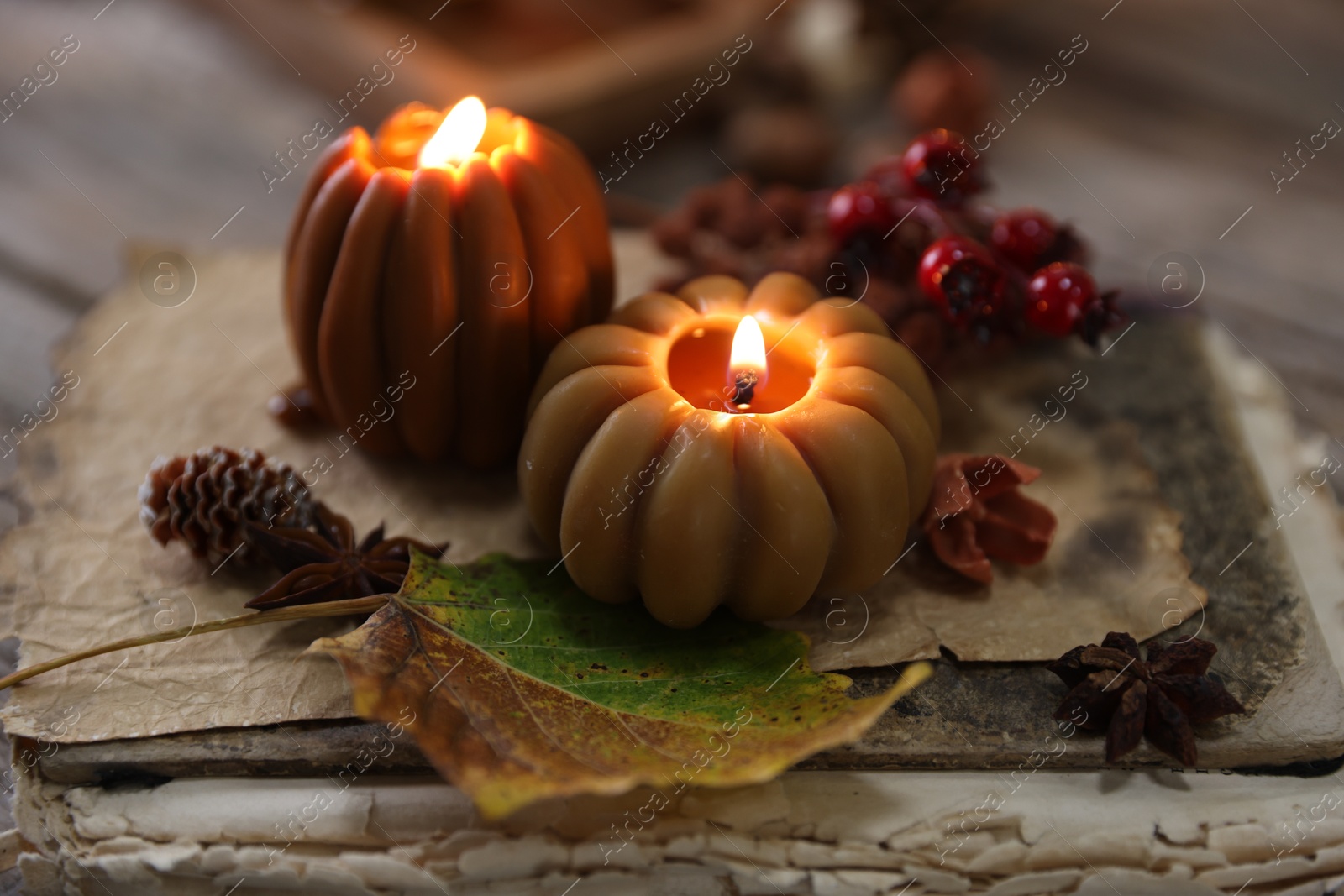 Photo of Burning candles in shape of pumpkins, old book and autumn decor on table, closeup