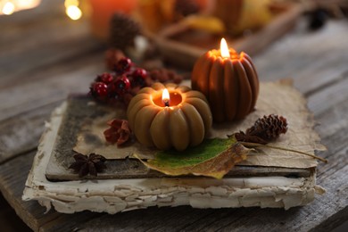 Photo of Burning candles in shape of pumpkins, old book and autumn decor on wooden table, closeup