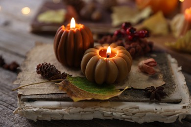 Burning candles in shape of pumpkins, old book and autumn decor on wooden table, closeup