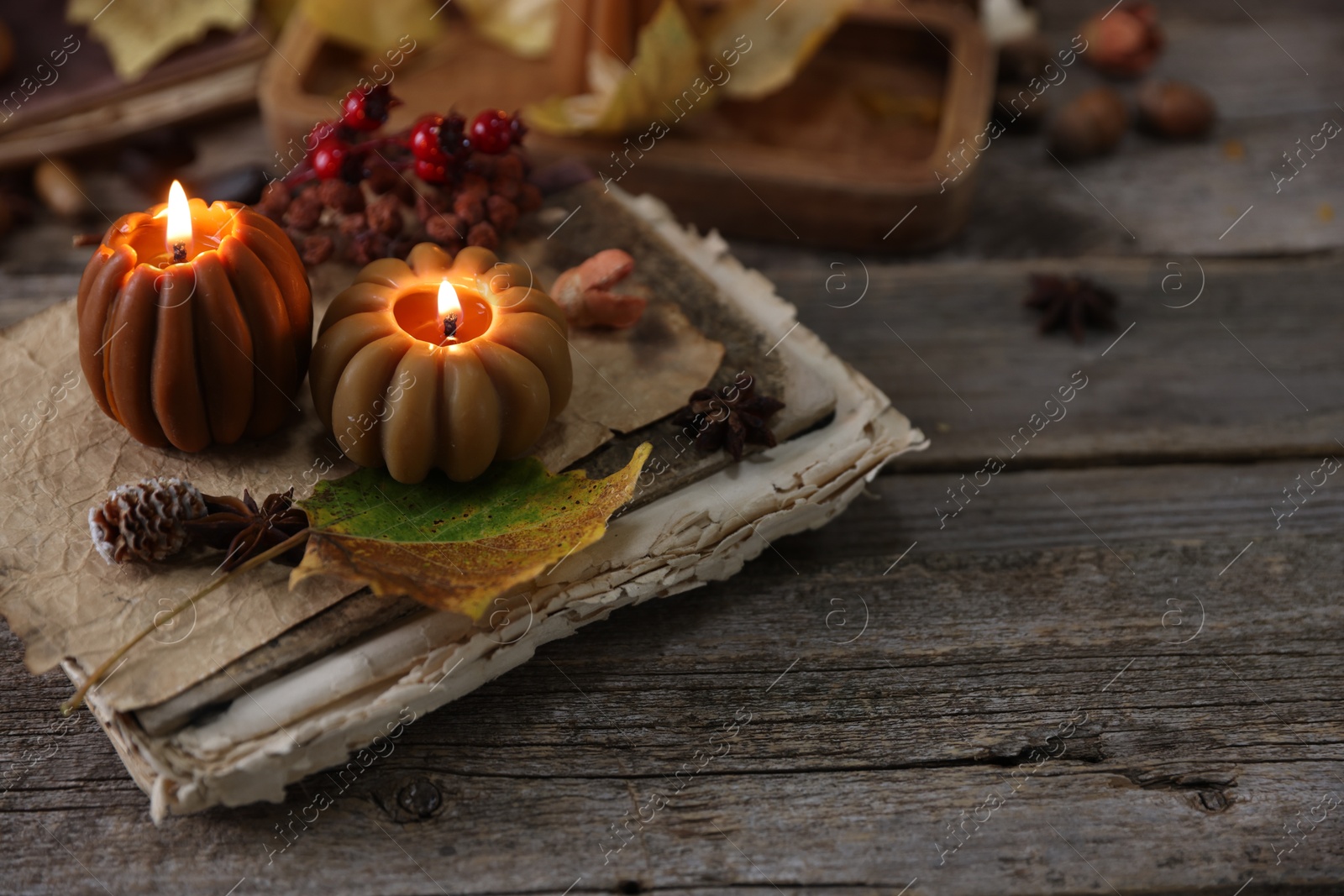 Photo of Burning candles in shape of pumpkins, old book and autumn decor on wooden table, closeup