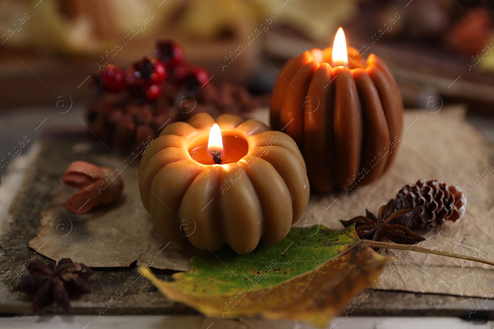 Photo of Burning candles in shape of pumpkins, old book and autumn decor on table, closeup