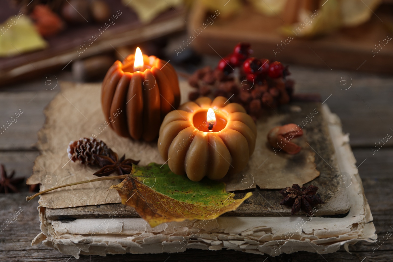 Photo of Burning candles in shape of pumpkins, old book and autumn decor on wooden table, closeup