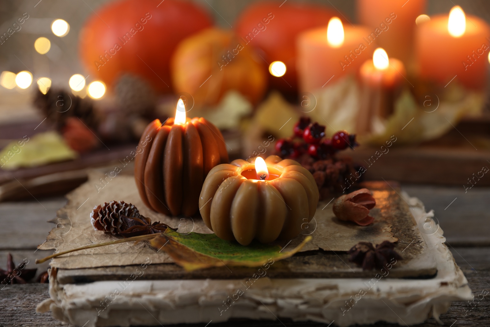 Photo of Burning candles in shape of pumpkins, old book and autumn decor on wooden table, closeup