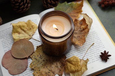 Photo of Burning candle, dry leaves and open book on green background, closeup. Autumn atmosphere