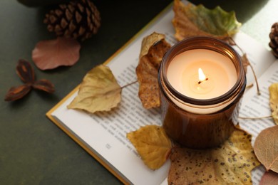 Photo of Burning candle, dry leaves and open book on green background, closeup. Autumn atmosphere