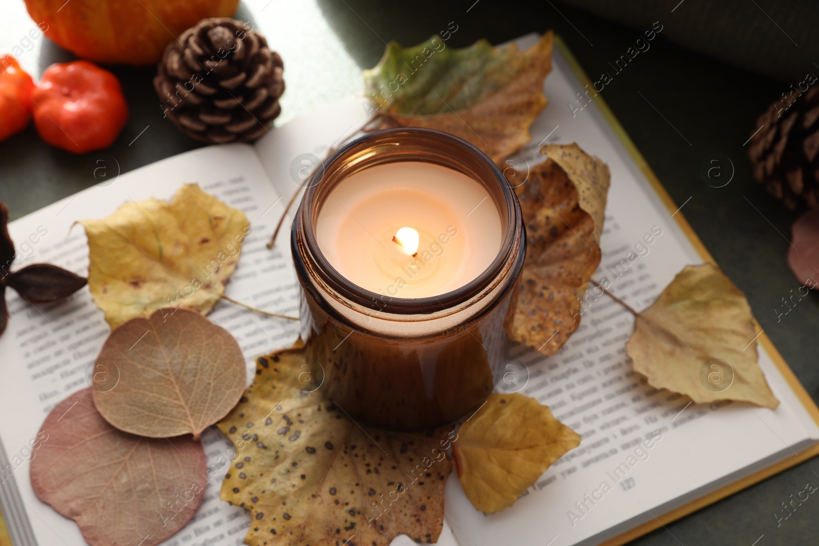 Photo of Burning candle, dry leaves and open book on green background, closeup. Autumn atmosphere