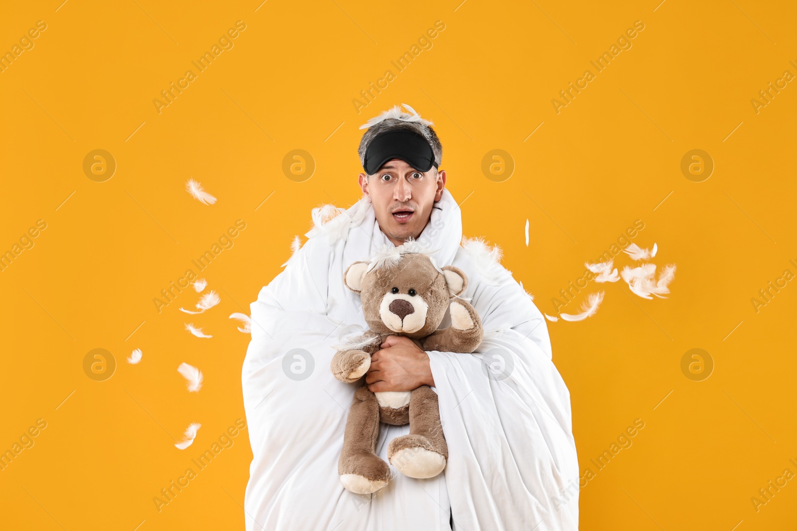 Photo of Overslept man with sleep mask and teddy bear wrapped in blanket on orange background