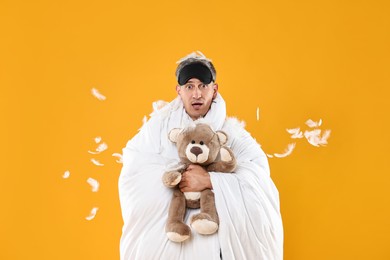 Photo of Overslept man with sleep mask and teddy bear wrapped in blanket on orange background