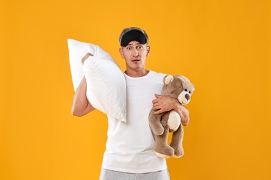 Photo of Overslept man with sleep mask, pillow and teddy bear on orange background