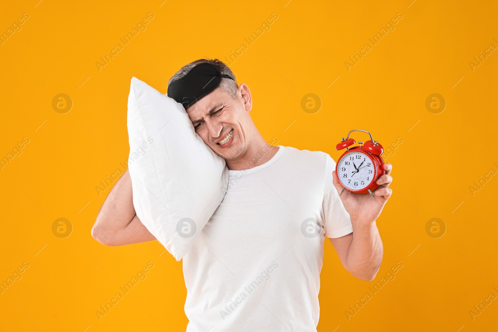 Photo of Overslept man with sleep mask, pillow and alarm clock on orange background