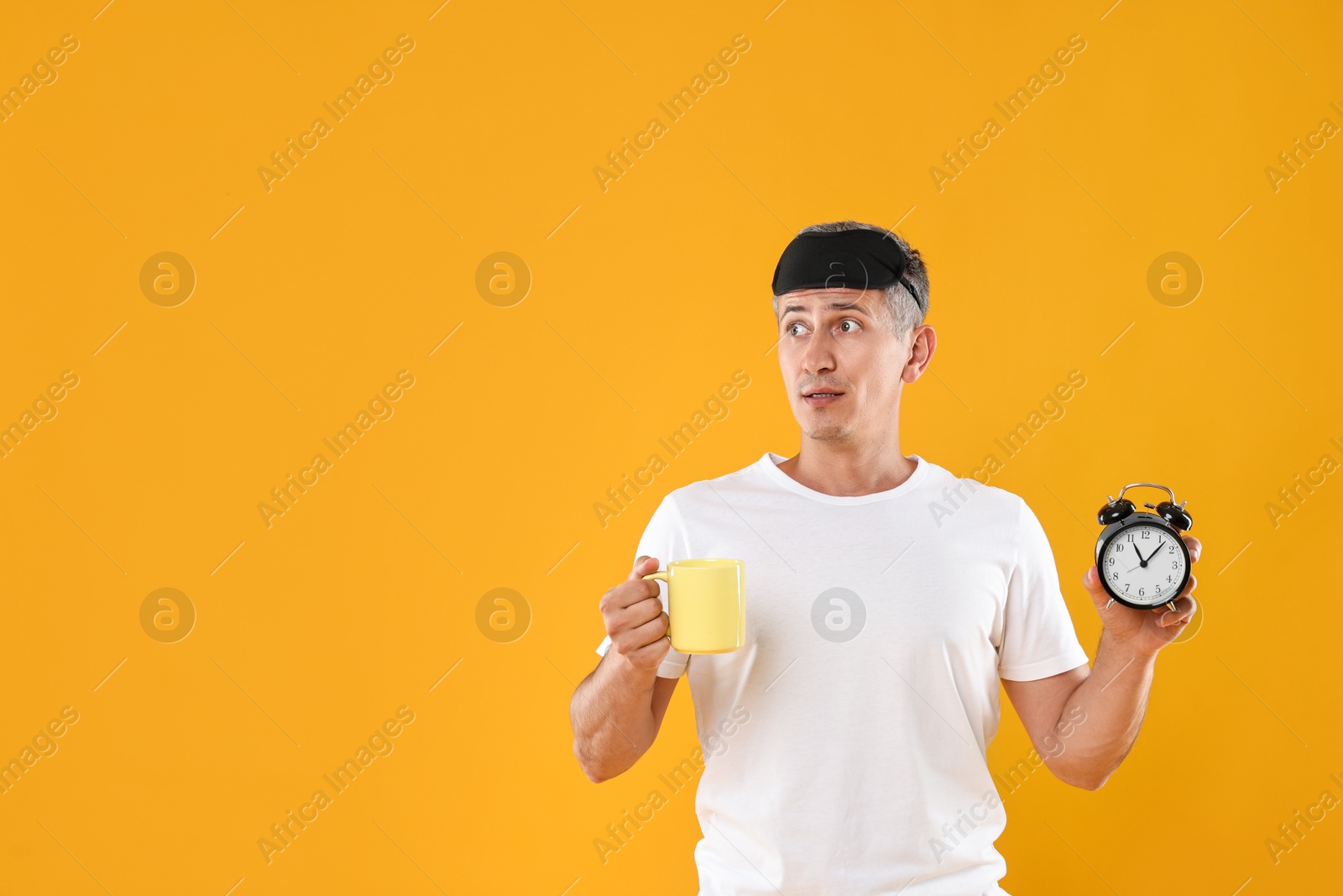 Photo of Overslept man with sleep mask, cup and alarm clock on orange background, space for text