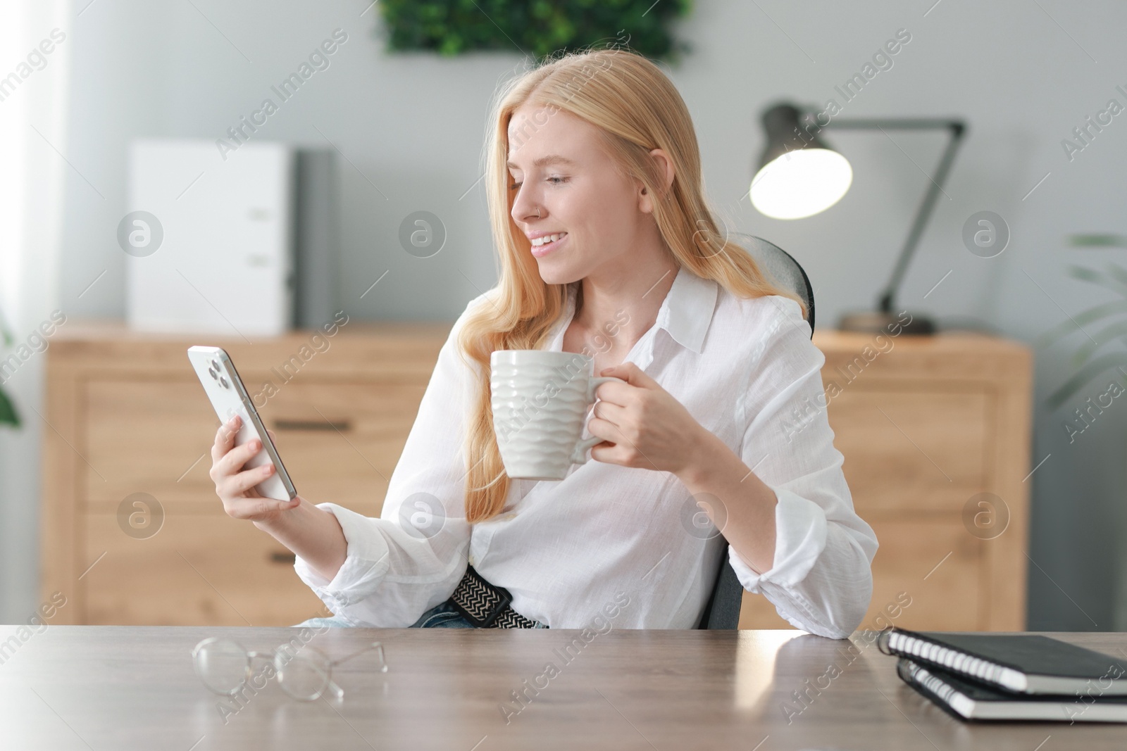 Photo of Smiling woman using smartphone and drinking coffee at table in office