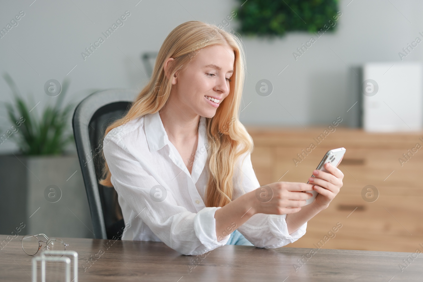 Photo of Smiling woman using smartphone and drinking coffee at table in office