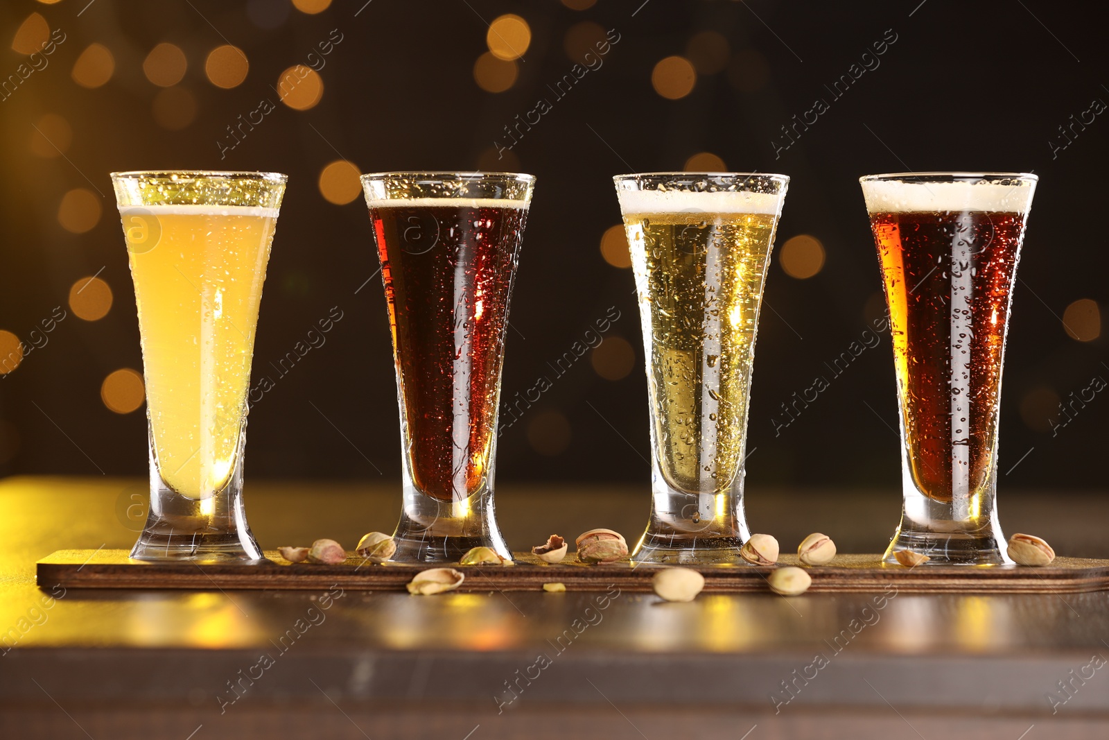 Photo of Glasses with different types of beer and pistachios on wooden table against blurred lights, closeup