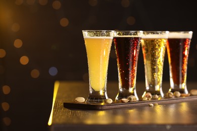 Photo of Glasses with different types of beer and pistachios on wooden table against blurred lights, closeup. Space for text