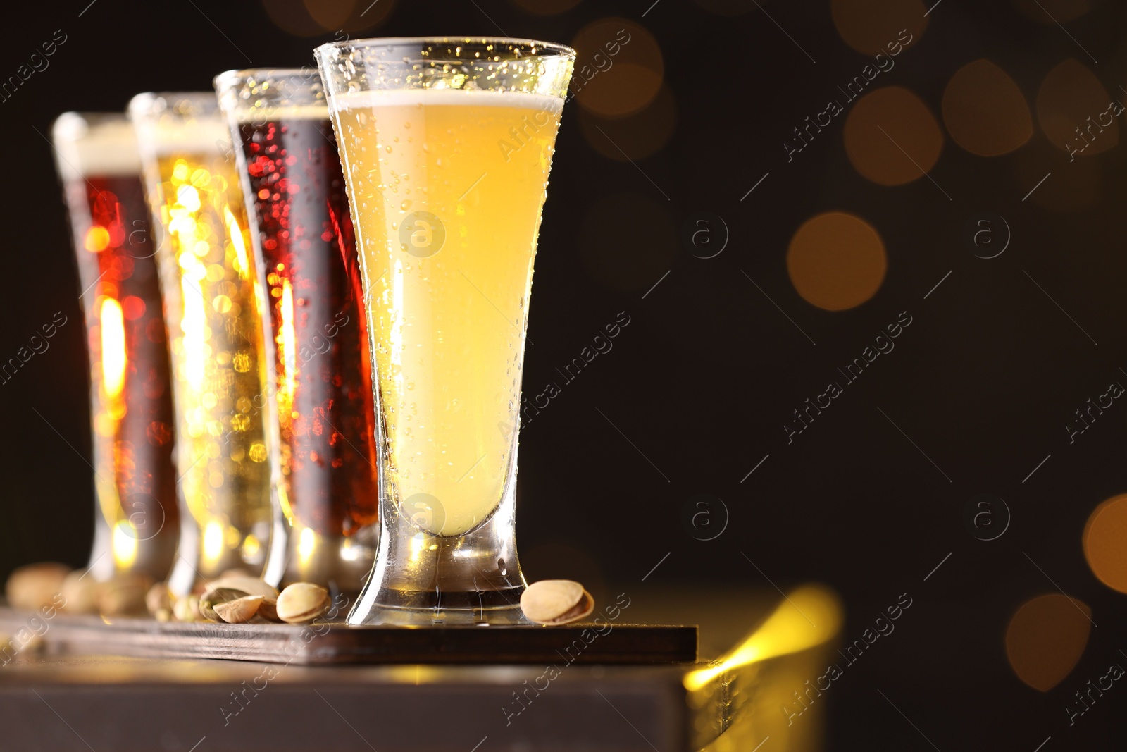 Photo of Glasses with different types of beer and pistachios on wooden table against blurred lights, closeup. Space for text