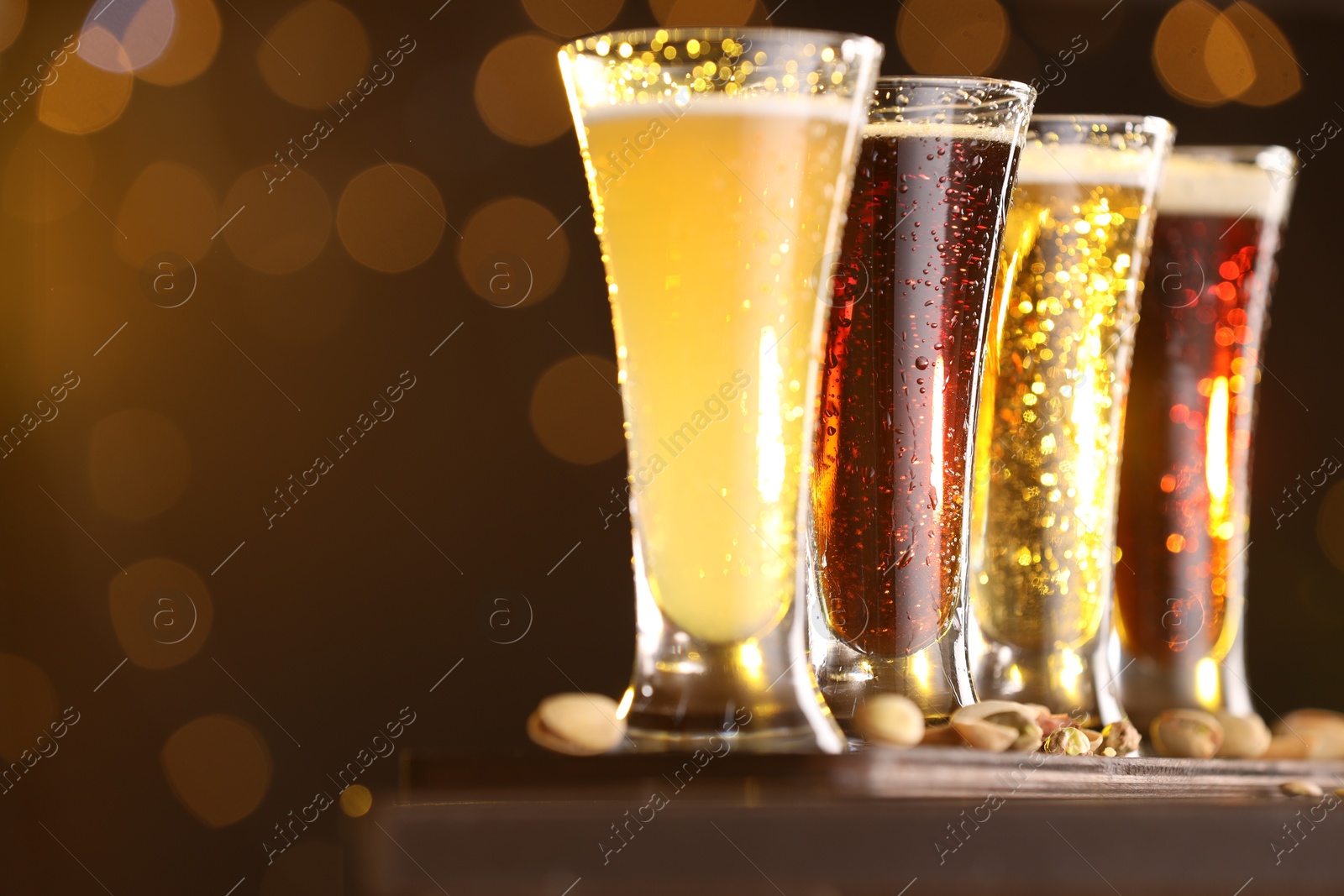 Photo of Glasses with different types of beer and pistachios on wooden table against blurred lights, closeup. Space for text