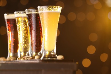 Photo of Glasses with different types of beer and pistachios on wooden table against blurred lights, closeup. Space for text