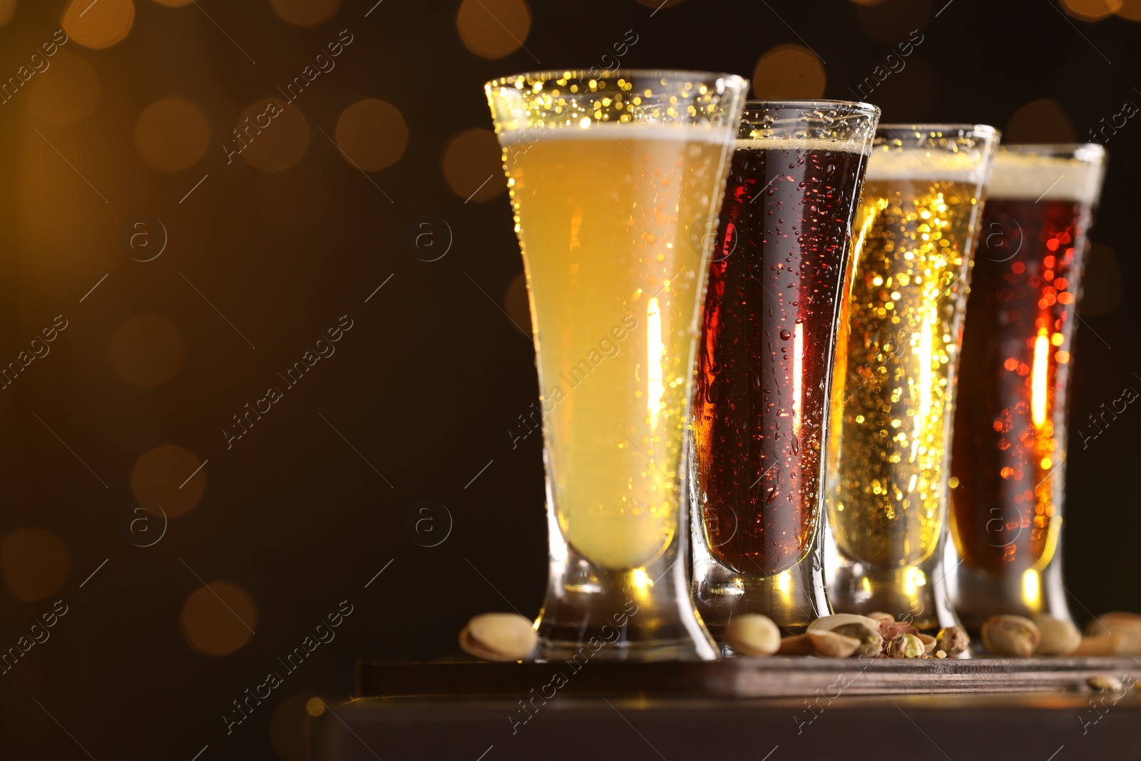 Photo of Glasses with different types of beer and pistachios on wooden table against blurred lights, closeup. Space for text