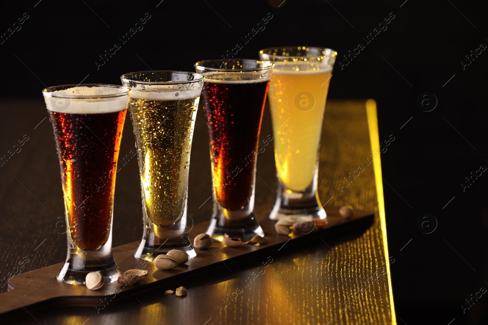 Photo of Glasses with different types of beer and pistachios on wooden table against black background, closeup