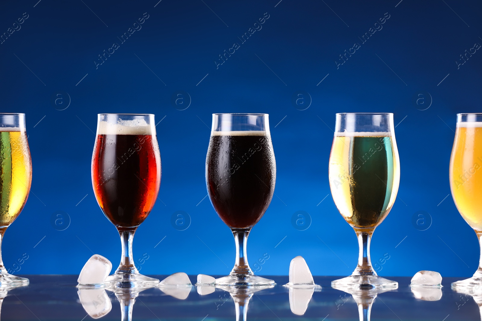 Photo of Glasses with different types of beer and ice cubes on mirror surface against blue background