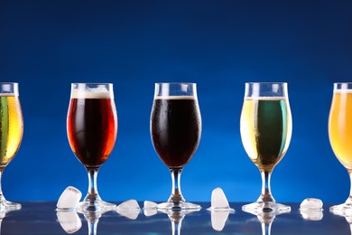 Photo of Glasses with different types of beer and ice cubes on mirror surface against blue background