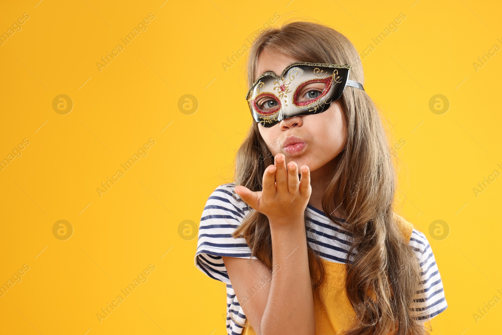 Photo of Cute girl wearing carnival mask and blowing kiss on orange background, space for text