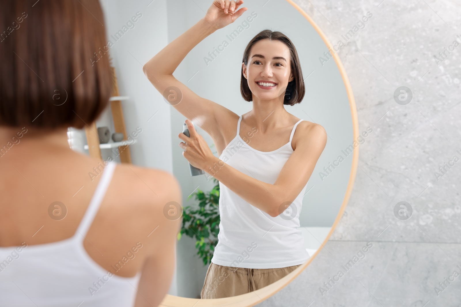 Photo of Smiling woman applying spray deodorant near mirror at home