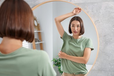 Photo of Emotional woman in t-shirt before using deodorant near mirror at home