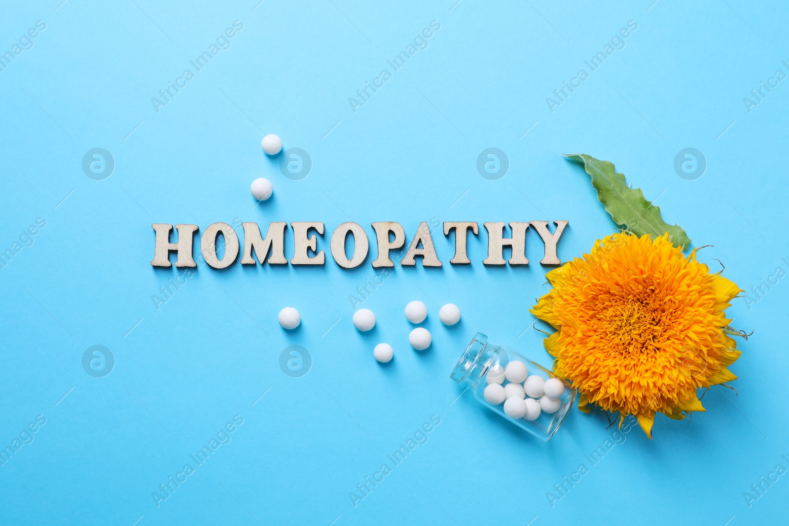 Photo of Word Homeopathy, pills and flower on light blue background, flat lay