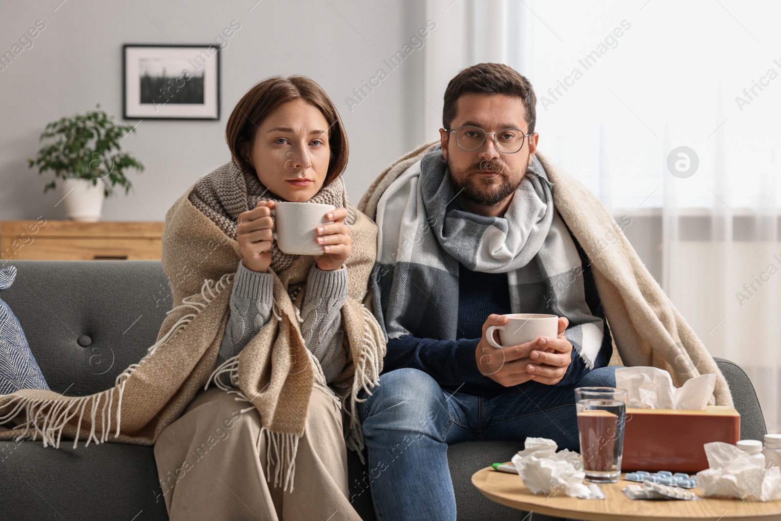 Photo of Cold symptom. Couple with cups of drink suffering from fever on sofa at home