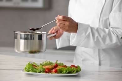 Professional chef adding sauce to dish with baked chicken at white marble table in kitchen, closeup