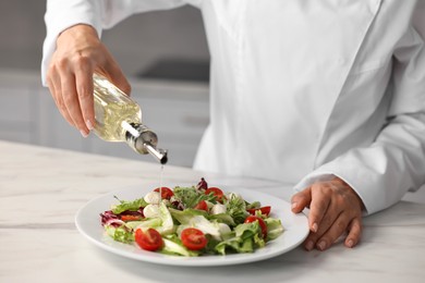 Photo of Professional chef pouring oil onto delicious salad at white marble table in kitchen, closeup