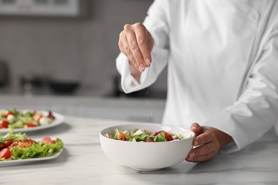 Professional chef adding salt to delicious salad at white marble table in kitchen, closeup