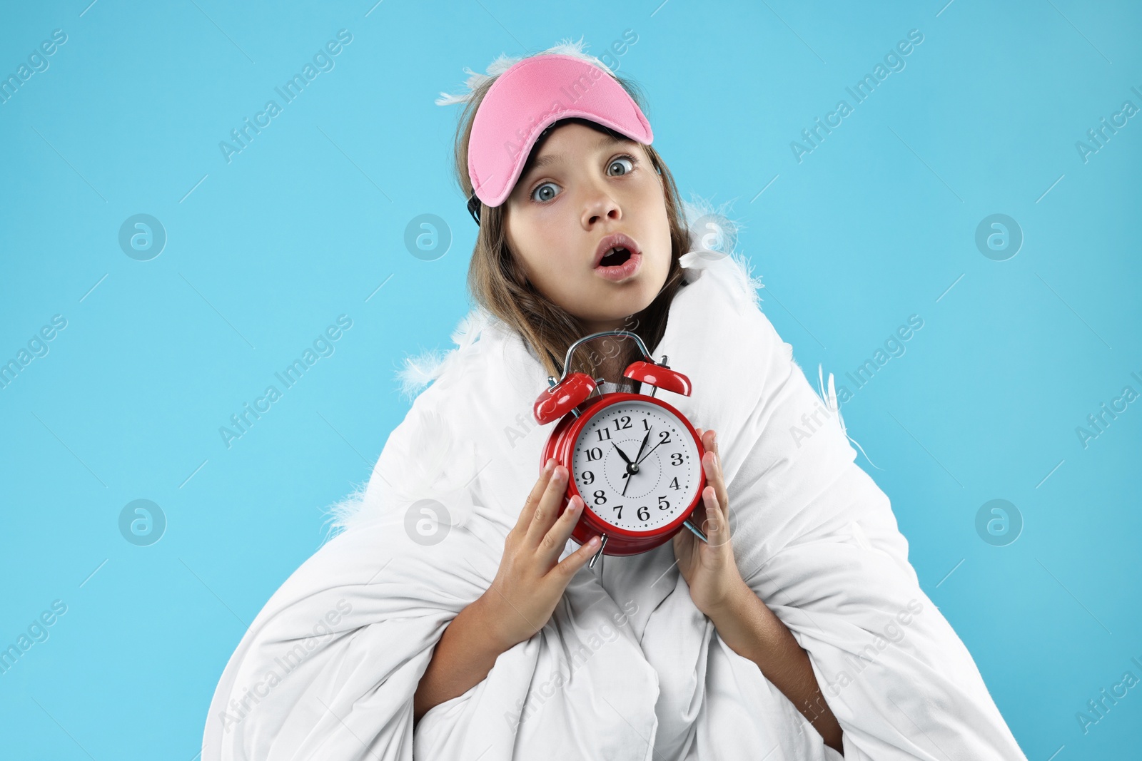 Photo of Overslept girl with sleep mask and alarm clock wrapped in blanket on light blue background