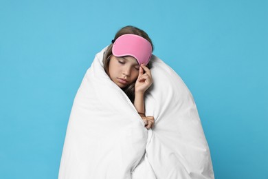 Photo of Overslept girl with sleep mask wrapped in blanket on light blue background