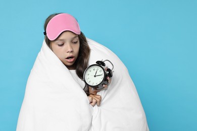 Photo of Overslept girl with sleep mask and alarm clock wrapped in blanket on light blue background, space for text