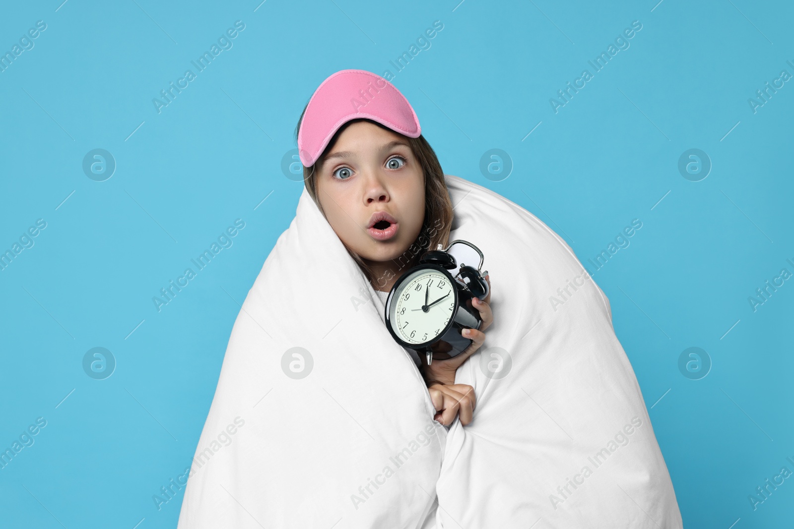 Photo of Overslept girl with sleep mask and alarm clock wrapped in blanket on light blue background