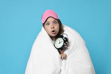 Photo of Overslept girl with sleep mask and alarm clock wrapped in blanket on light blue background